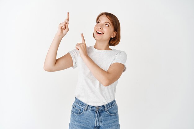 Señora bonita emocionada en camiseta apuntando con el dedo hacia arriba, sonriendo y mirando divertido, mirando publicidad de promoción, de pie en la pared blanca