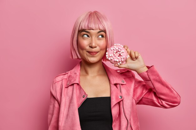 Señora bonita de ensueño con el pelo teñido de bob, sostiene una deliciosa y apetitosa dona tiene la tentación de comer un postre alto en calorías, usa una elegante chaqueta rosa