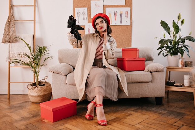 Señora bonita en gabardina posa pensativamente y sostiene los zapatos. Mujer bonita en traje elegante se sienta en un acogedor sofá con tacones rojos.
