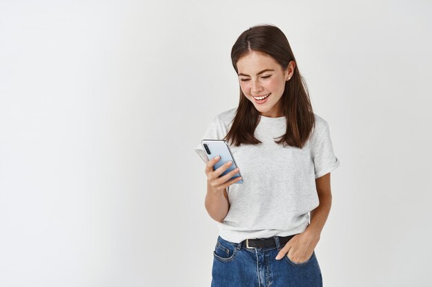 Señora bonita joven leyendo un mensaje en el teléfono inteligente, mirando la pantalla del móvil y sonriendo, de pie en ropa casual sobre una pared blanca