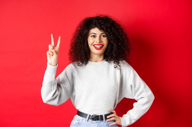 Señora bonita joven en sudadera que muestra el número dos, haciendo un pedido y sonriendo, de pie sobre fondo rojo.