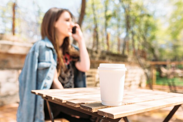 Señora bonita joven vestida con chaqueta de mezclilla está hablando por teléfono en el parque en la luz del sol con una sonrisa encantadora. Al primer plano una taza con café. Día soleado, buen humor.