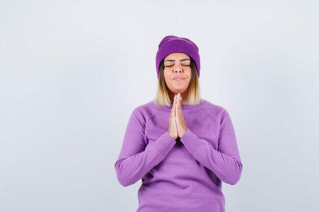 Señora bonita con las manos en gesto de oración en suéter, gorro y mirando esperanzado, vista frontal.