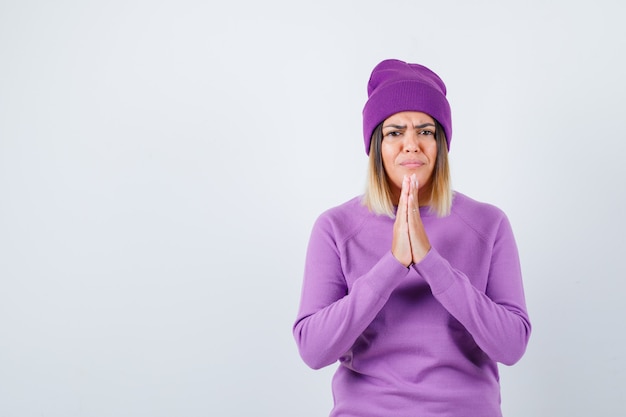 Señora bonita con las manos en gesto de oración en suéter, gorro y mirando triste. vista frontal.