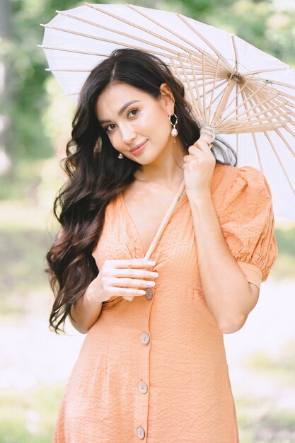 Señora bonita que sostiene el paraguas y que mira en naturaleza en vestido anaranjado durante día.