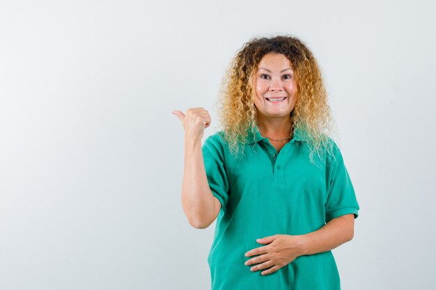 Señora bonita rubia apuntando hacia atrás con el pulgar en la camiseta de polo verde y mirando alegre. vista frontal.