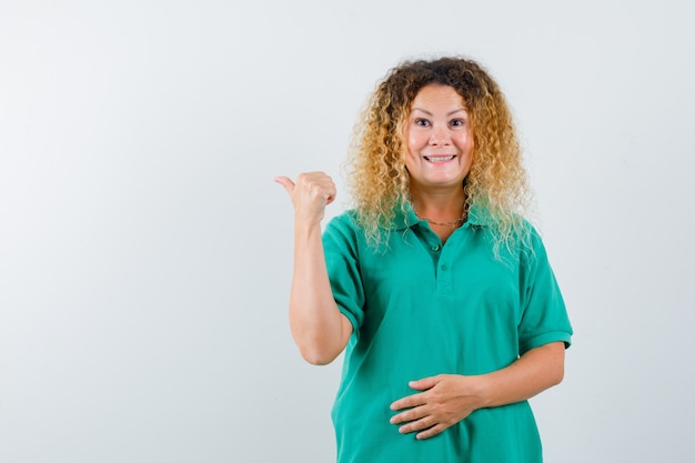 Señora bonita rubia apuntando hacia atrás con el pulgar en la camiseta de polo verde y mirando alegre. vista frontal.