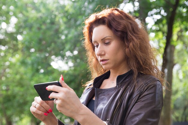 Señora bonita seria navegando en el teléfono inteligente en el parque de la ciudad