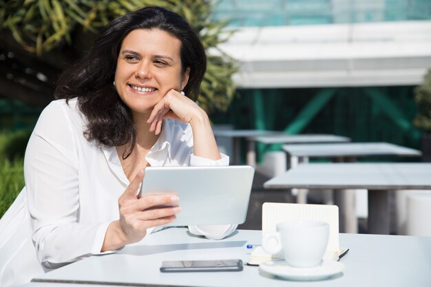 Señora bonita sonriente que trabaja y que usa la tableta en café de la calle