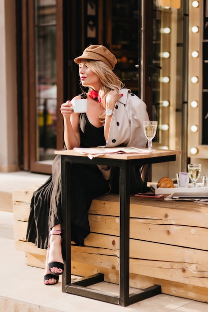 Señora bonita en vestido largo y sandalias negras disfrutando del almuerzo en la cafetería al aire libre y mirando a otro lado. Fascinante chica rubia con sombrero esperando amigo para comer croissants juntos.