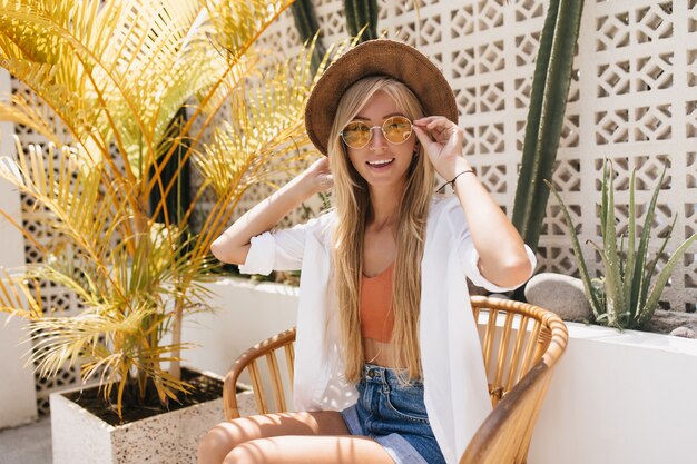 Señora bronceada inspirada en elegante sombrero escalofriante en la cafetería del complejo por la mañana. Atractiva turista pasar el fin de semana al aire libre.