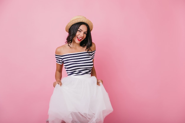 Señora bronceada segura que presenta en elegante sombrero de paja. Retrato de niña riendo jocund jugando con falda blanca.