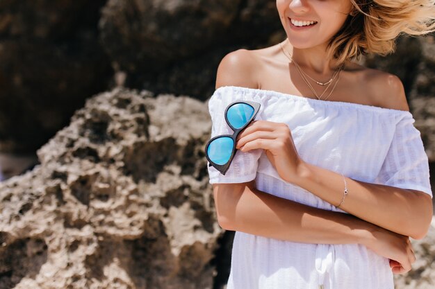 Señora bronceada en vestido y pulsera posando junto a la roca. Retrato al aire libre de mujer elegante con gafas de sol.