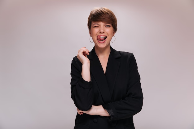 Señora de buen humor posando sobre fondo aislado y mostrando la lengua. Mujer bonita en chaqueta negra sonríe sobre fondo blanco