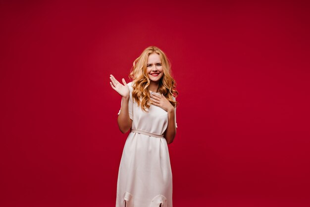 Señora de buen humor soñadora posando en vestido blanco. extática chica de pelo largo aislada en la pared roja.