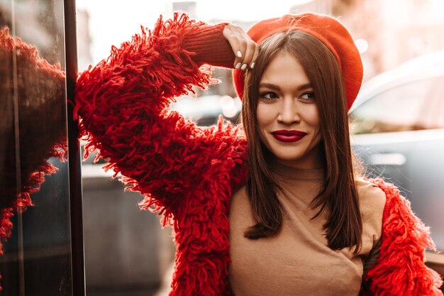 Señora con cabello lacio oscuro y ojos marrones vestida con boina roja y abrigo ecológico posando en la calle, apoyada en la pared de vidrio.