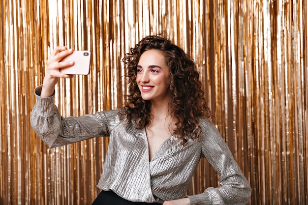 Señora de cabello oscuro en blusa plateada tomando selfie sobre fondo dorado