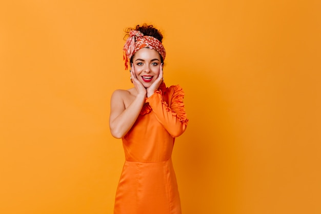 Señora de cabello oscuro con lápiz labial rojo vestida con vestido naranja y banda delantera con una sonrisa mirando a la cámara en el espacio aislado.