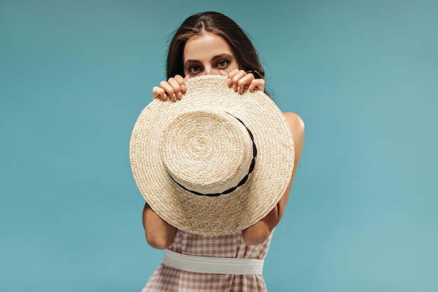 Foto gratuita señora de cabello oscuro en vestido de verano rosa con cinturón ancho que cubre su rostro con elegante sombrero de ala ancha sobre fondo azul