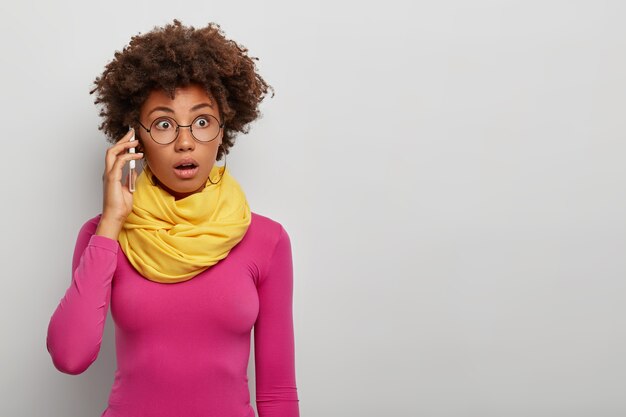 La señora de cabello rizado horrorizada telefonea a un colega, usa un teléfono celular moderno, se concentra en obtener información durante la conversación telefónica, usa gafas