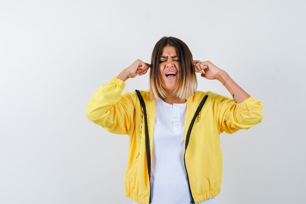 Señora en camiseta, chaqueta tapando las orejas con los dedos y mirando molesta, vista frontal.