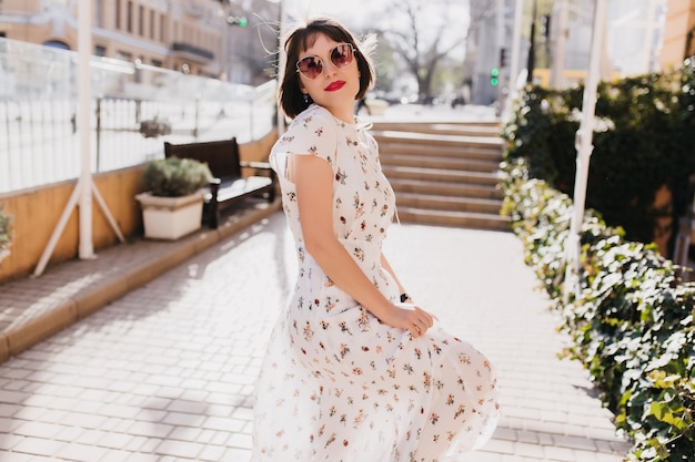 Señora caucásica alegre en gafas de sol negras caminando por la ciudad en día de verano. Tiro al aire libre de chica interesada de pelo corto en vestido largo blanco escalofriante en mañana soleada de primavera.