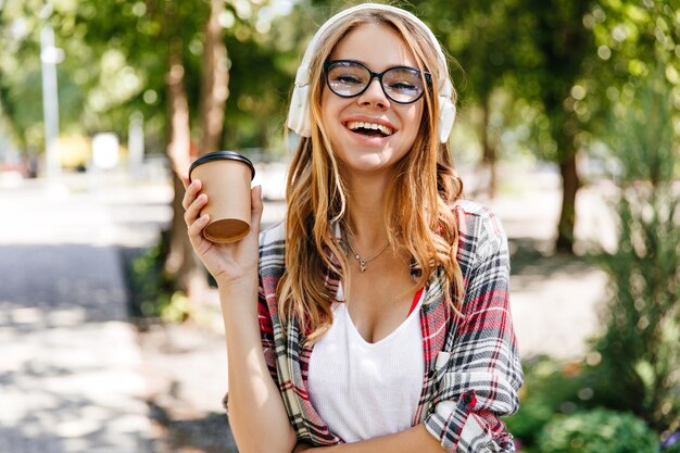 Señora caucásica elegante expresando emociones positivas en el parque. Foto al aire libre de la hermosa mujer sonriente tomando café en la naturaleza.