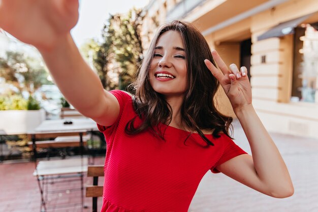 Señora caucásica entusiasta haciendo selfie en café al aire libre. Riendo mujer elegante tomando una foto de sí misma.