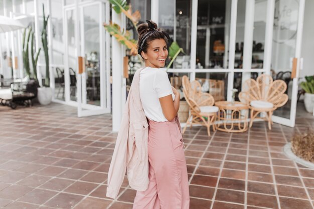 Señora en chaqueta y pantalones de mezclilla rosa está sonriendo afablemente mientras pasa por street cafe