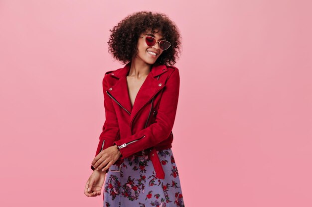 Señora en chaqueta roja y falda con sonrisa posando sobre fondo rosa Retrato interior de mujer con gafas de sol sonriendo sobre fondo aislado