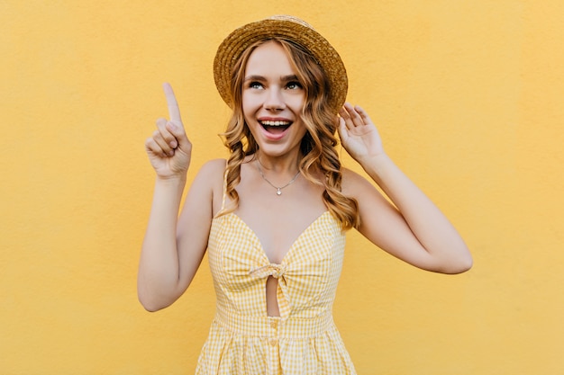 Señora delgada inspirada mirando hacia arriba con sonrisa alegre. Retrato interior de una hermosa niña emocional viste un vestido amarillo.