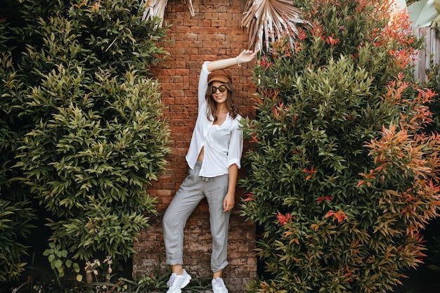 Señora delgada en zapatillas blancas, pantalones grises y blusa blanca de gran tamaño posa contra la pared de ladrillo rodeada de arbustos.