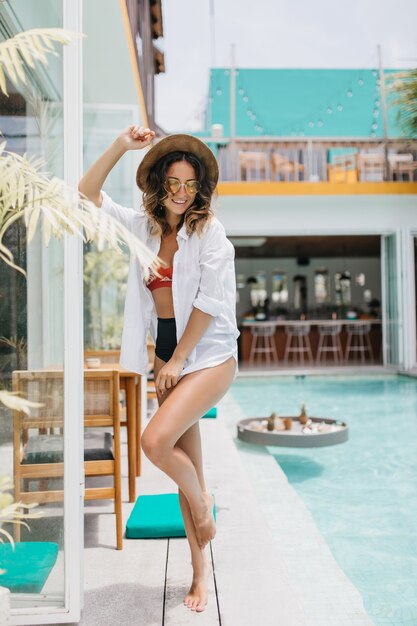 Señora descalza en camisa blanca descansando en el lugar de veraneo. mujer morena lleva sombrero bailando junto a la piscina.