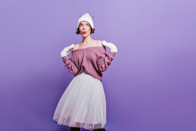 Señora despreocupada en guantes de lana posando con expresión de cara de ensueño. Retrato de interior de señorita con gorro de punto y falda larga blanca aislada en la pared púrpura.