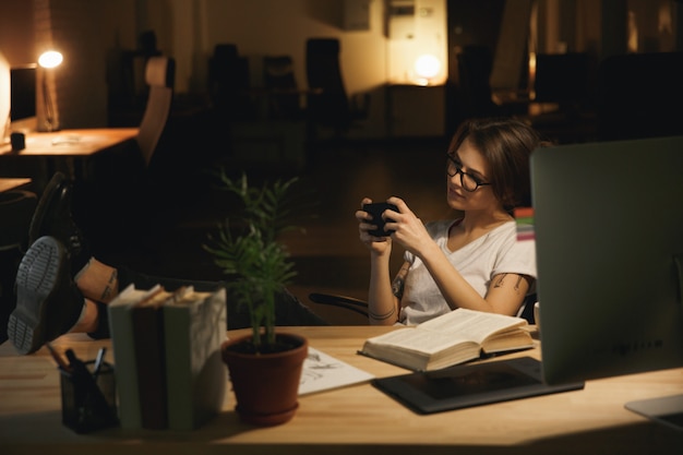 Señora diseñadora sentada en el interior por la noche jugar juegos por teléfono.