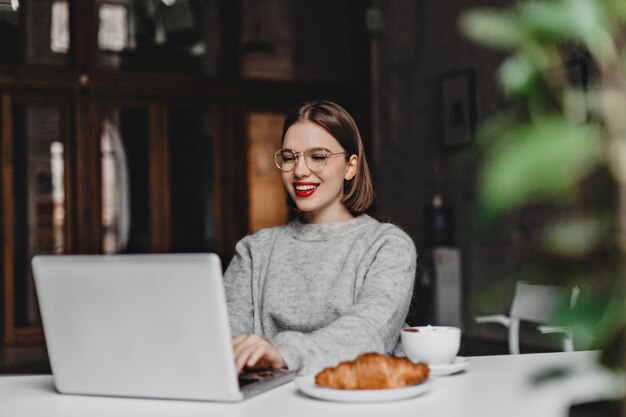 Señora elegante con gafas y suéter de cachemira con una sonrisa trabajando en un portátil gris, sentado en la cafetería con croissant y una taza de café en la mesa.