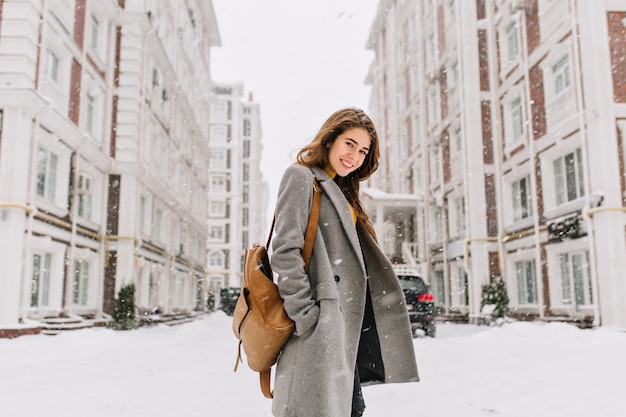 Señora elegante con mochila marrón caminando por la ciudad bajo la nieve. Foto al aire libre de una mujer bonita con una sonrisa encantadora posando en abrigo gris en la escena urbana