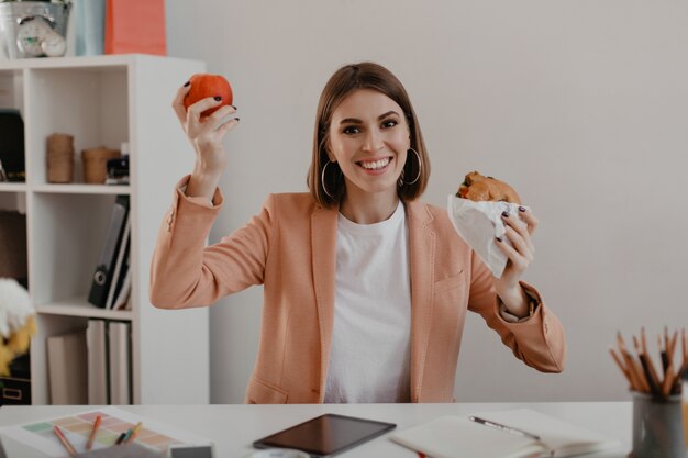 Señora con elegante traje ligero muestra su almuerzo que consiste en manzana y hamburguesa en el lugar de trabajo.