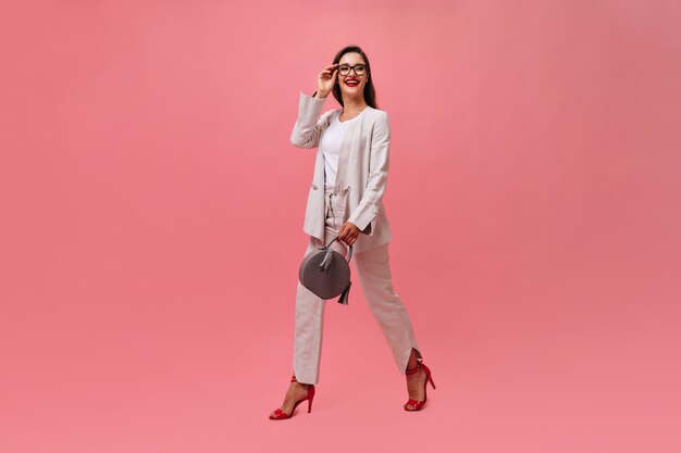 Señora elegante en traje sostiene el bolso y camina sobre fondo rosa. Mujer de negocios con cabello oscuro con labios rojos brillantes y elegantes tacones sonriendo.