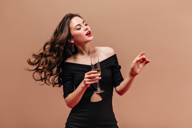 Foto gratuita señora elegante en vestido negro con copa de vino espumoso sobre fondo beige.