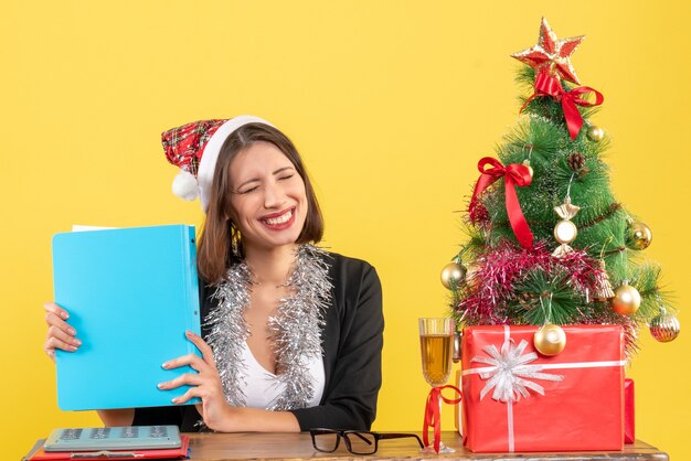 Señora encantadora feliz satisfecha en traje con sombrero de santa claus y decoraciones de año nuevo con documento en la oficina en amarillo aislado