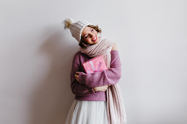 Señora entusiasta en falda blanca y gorro de punto posando con caja presente. Retrato interior de niña inspirada preparándose para el año nuevo y sosteniendo un regalo.