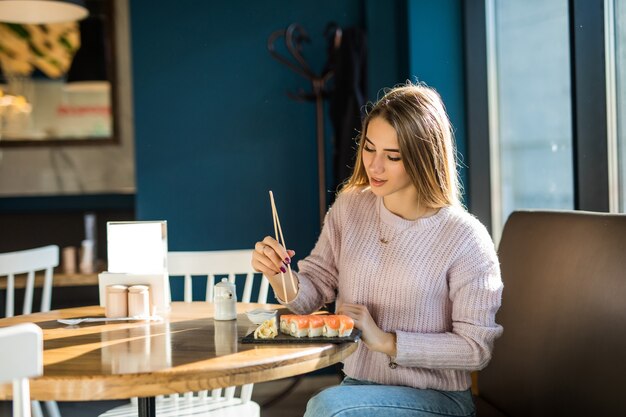 Señora estudiante en suéter blanco comiendo sushi para el almuerzo en una pequeña cafetería