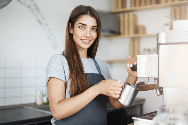 Señora experimentada barista desnatar la leche en una jarra mirando a la cámara sonriendo