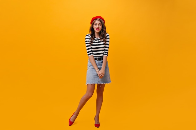 Señora en falda de mezclilla y boina roja posa sobre fondo naranja. Mujer joven en ropa de moda y tacones brillantes mirando a cámara.