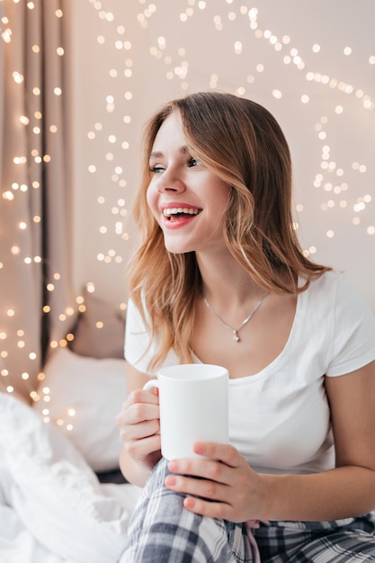 Señora fascinante en pijama tomando café. Retrato interior de alegre mujer rubia divirtiéndose en la mañana.