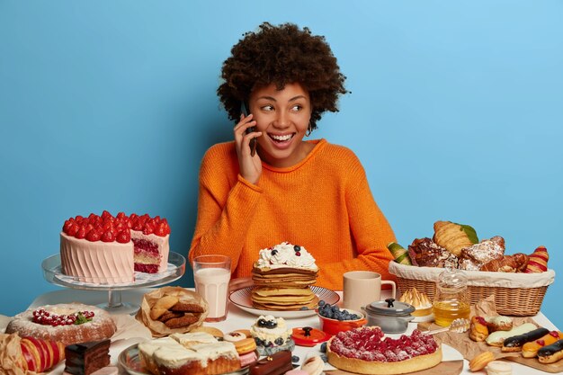 Señora feliz con corte de pelo afro tiene una conversación agradable a través del teléfono celular, disfruta comiendo pasteles sabrosos, considera comer panqueques con crema, siendo goloso, aislado en la pared azul.