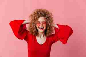 Foto gratuita señora feliz despeina su cabello rubio rizado. chica con gafas y blusa brillante se ríe en el espacio rosa.
