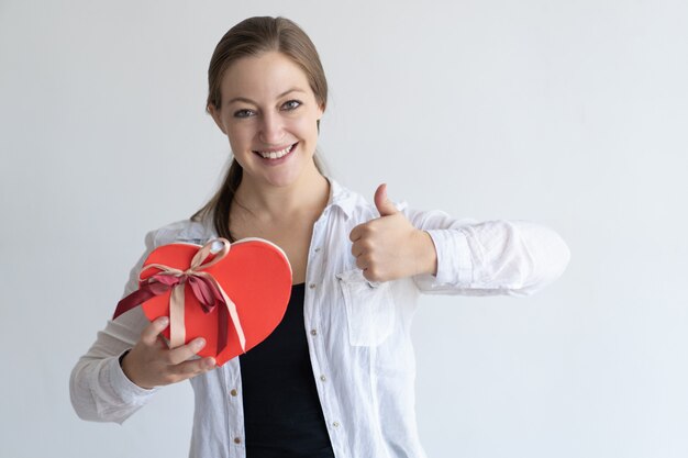 Señora feliz que muestra la caja y el pulgar en forma de corazón de regalo para arriba