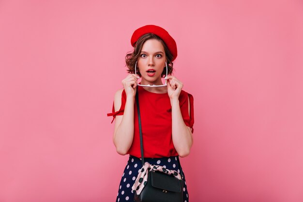 Señora francesa romántica posando con expresión de la cara de sorpresa. Filmación en interiores de adorable mujer rizada en boina roja aislada.